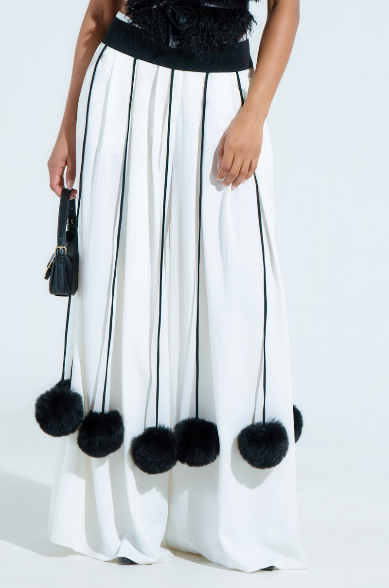 A person wearing a white skirt adorned with SHAKE YA POM POMS FUR BELT in black stripes ending with faux fur pom poms holds a small black purse in their left hand against a plain background.