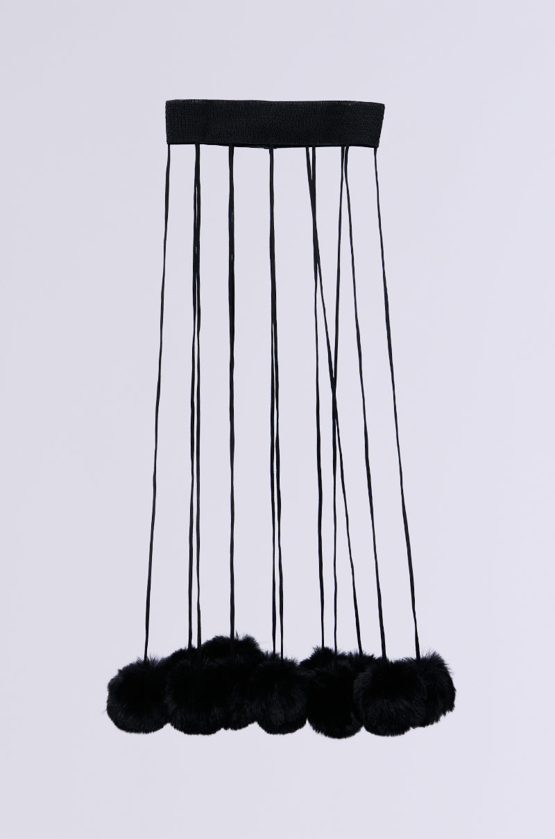 The SHAKE YA POM POMS FUR BELT is a modern black belt featuring fringe attachments and long elastic strings ending in faux fur pom-poms, set against a plain light gray background.
