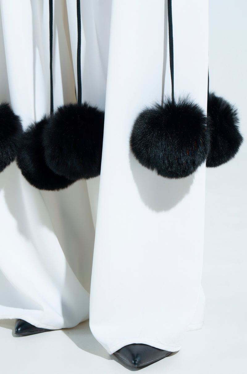 A close-up showcases the SHAKE YA POM POMS FUR BELT, featuring fringe details and black faux fur pompoms along the hem, draping over pointed black shoes and casting a soft shadow on the light surface.