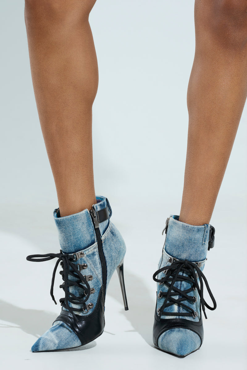 Close-up of legs showcasing the AZALEA WANG BLANCHE DENIM STILETTO BOOTIE with a pointed toe, ankle-high shaft, black accents, laces, side zippers, and metal eyelets against a plain background.
