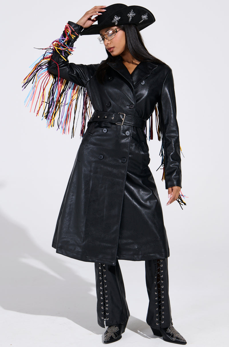Someone wearing the RYDER FRINGE TRENCH IN BLACK MULTI with colorful fringe sleeves, matching lace-up pants, a black cowboy hat with silver details, glasses, and pointed boots.