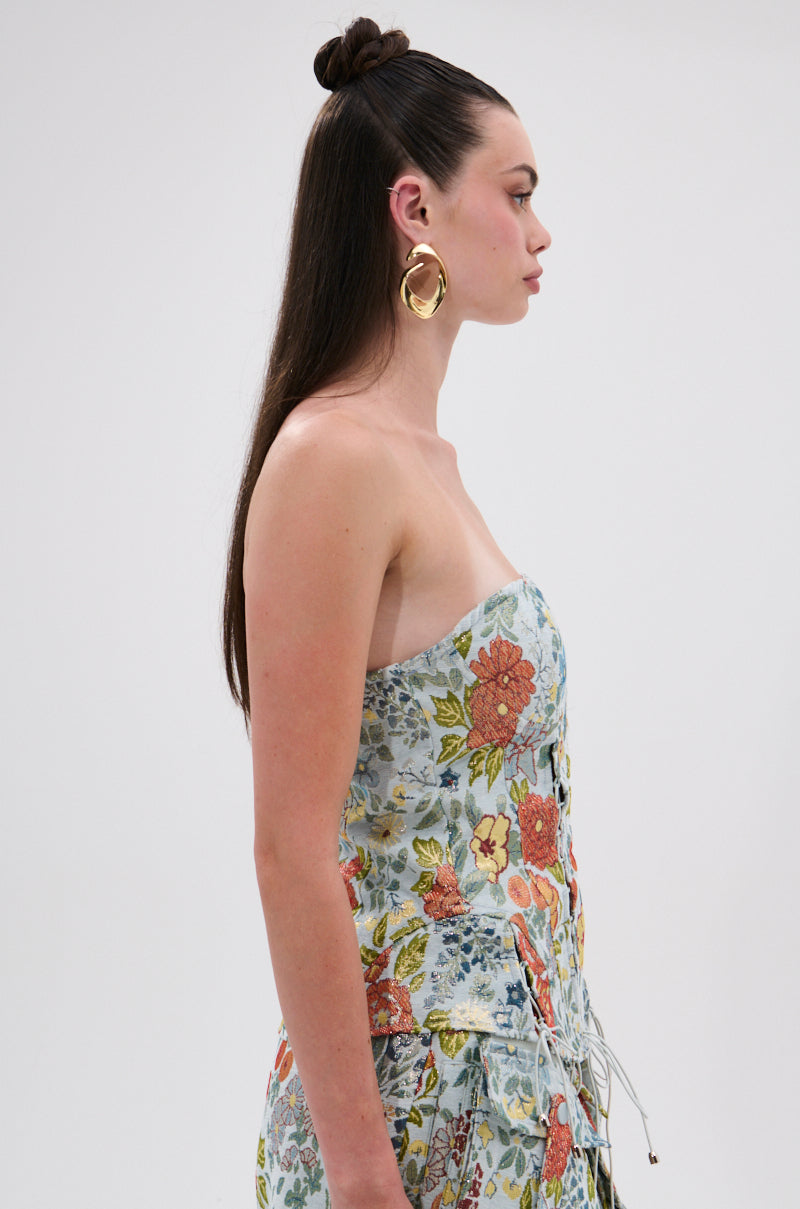 A woman with long, straight brown hair in a top knot wears the MARTHA FLORAL CORSET dress and large gold hoop earrings, facing sideways against a plain light background.