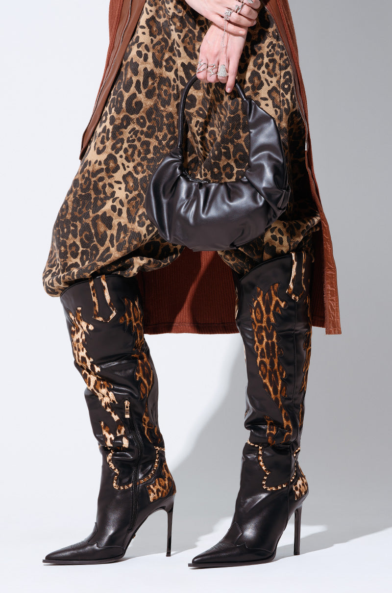 A person is wearing a brown, leopard-print outfit with AZALEA WANG BRIGGS LEOPARD BOOTS, featuring black, over-the-knee stiletto heels with faux leather upper and leopard print accents. They are holding a small, black handbag. Only the lower part of their body is visible.