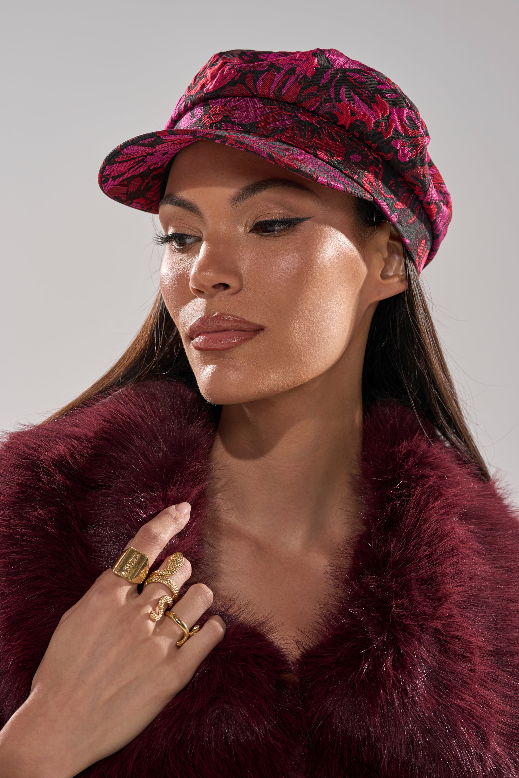 A woman wearing a textured burgundy faux fur coat and the LUXE LIVING BRIM HAT gazes to the side. She has long straight hair, bold winged eyeliner, and gold rings adorning her fingers.