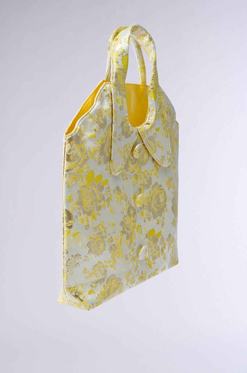 The COME WITH ME BROCADE PURSE is a rectangular tote bag with a floral brocade pattern in yellow and silver tones, featuring two short handles and a glossy finish for an elegant vintage-inspired look.