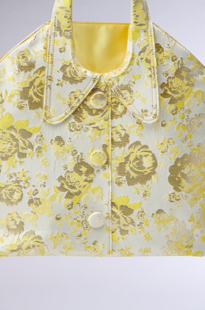 The COME WITH ME BROCADE PURSE is a vintage-inspired handbag like a collared shirt, featuring a yellow floral pattern on a pale background. It boasts two buttons, handles, and intricate stitching, making it an elegant and charming wardrobe accessory.