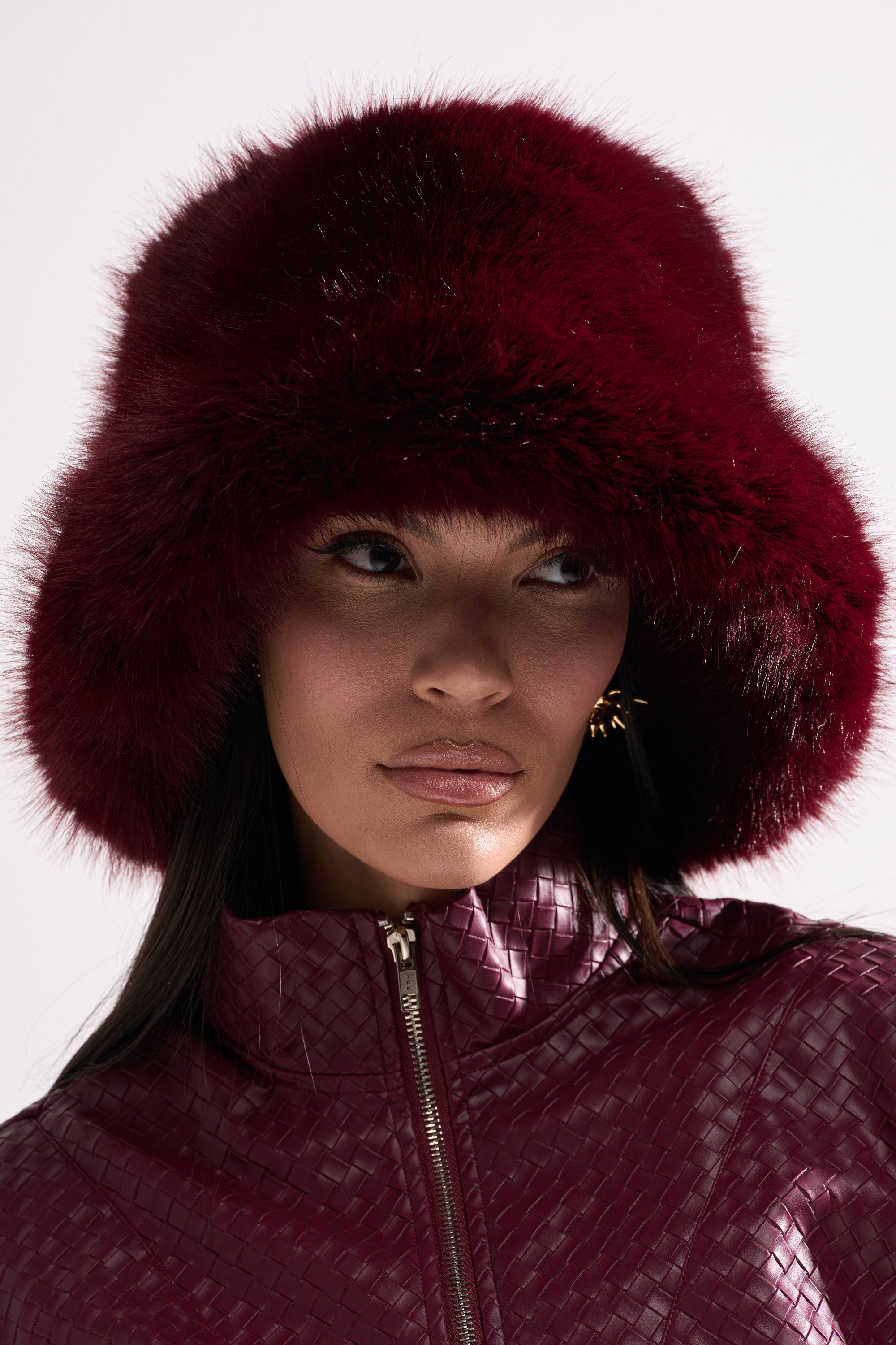 A woman wears the ELEVATED FAUX FUR BUCKET HAT in burgundy with a matching zip-up jacket, gazing slightly to the side with a neutral expression against a plain white background.