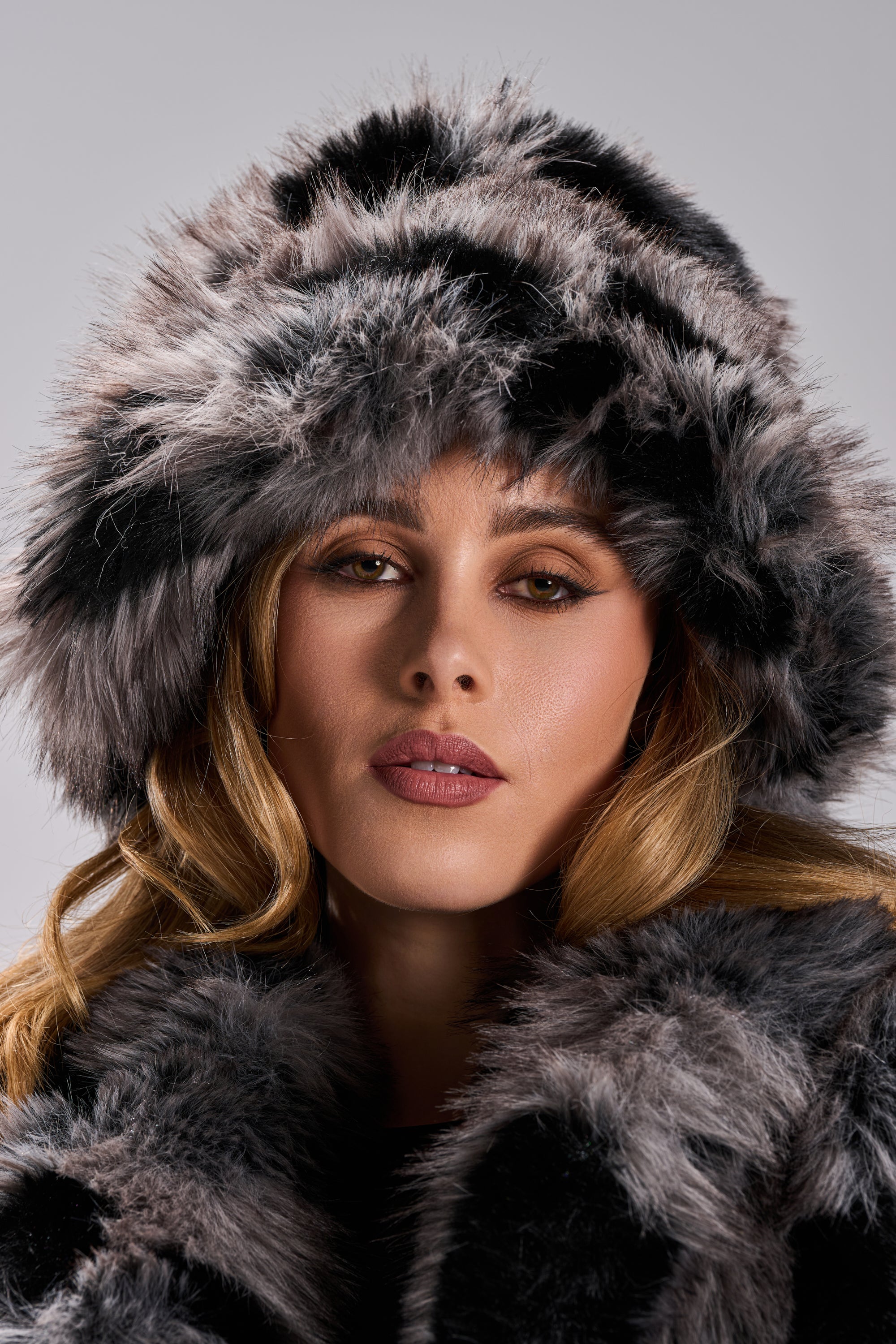A woman with wavy blonde hair wears the ARLO FAUX FUR BUCKET HAT and a matching coat, looking directly at the camera with a neutral expression against a plain gray background—a chic winter accessory statement.