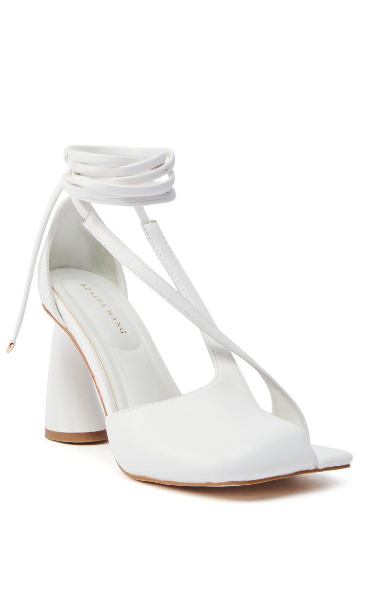 The AZALEA WANG BUIKY WHITE OPEN TOE PUMP is a white high-heeled sandal with wrap-around ankle straps, a sculpted chunky heel, and a modern minimalist design in faux leather. It features an open toe and a slightly cushioned insole for comfort.