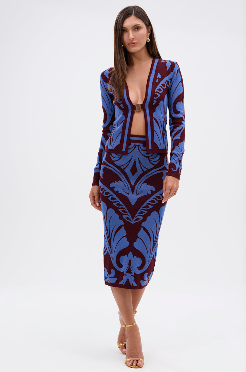 A woman stands against a white background wearing the GIVE ME LOVE MIDI SKIRT, featuring a deep red and cobalt blue damask knit with a pencil silhouette and high-rise waistband, paired with a matching open-front cardigan and strappy heels.