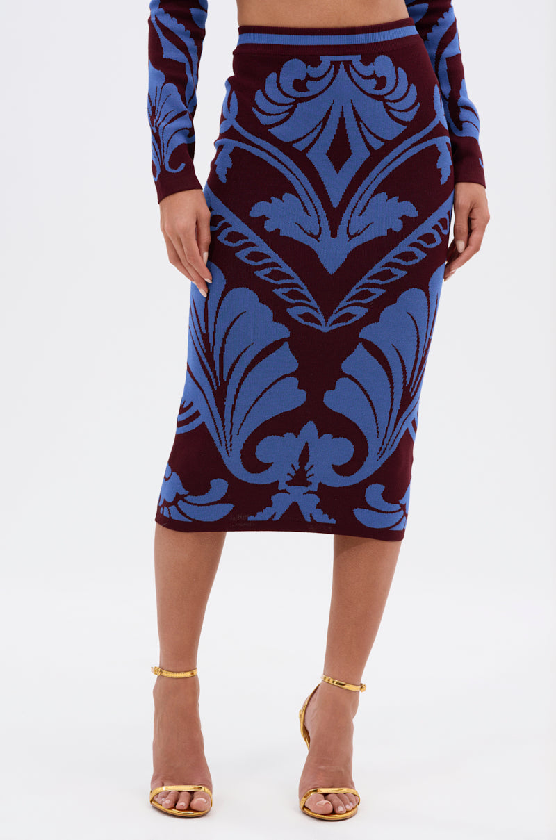 A woman wears the GIVE ME LOVE MIDI SKIRT, a fitted high-waisted pencil skirt in cobalt blue damask, paired with a matching long-sleeve top and gold strappy heels.
