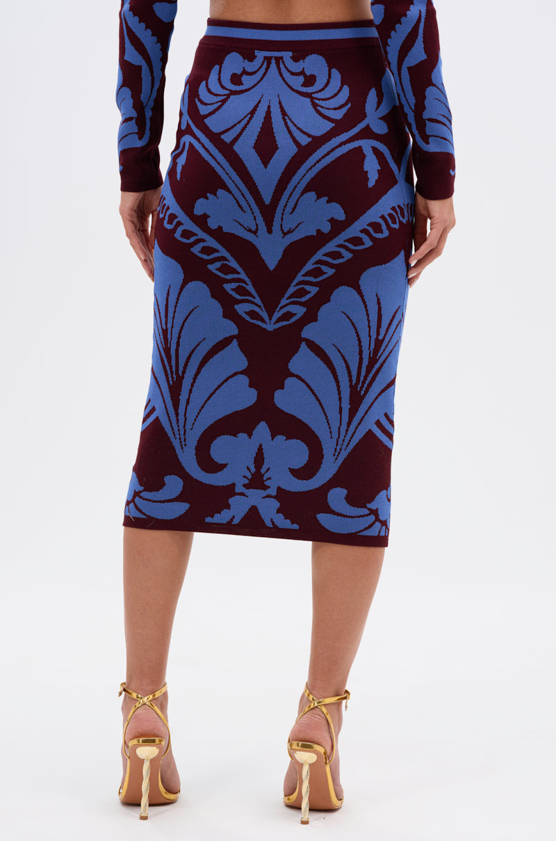 A woman faces away, wearing the GIVE ME LOVE MIDI SKIRT with a maroon and cobalt blue damask pattern, matching long sleeves, gold high-heeled sandals, a bold design, and a high-rise elastic waistband.