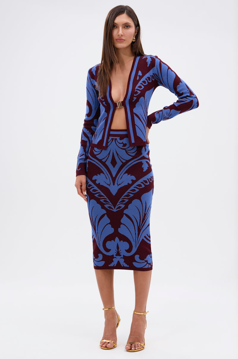 A woman faces forward in the GIVE ME LOVE MIDI SKIRT, a cobalt blue damask knee-length skirt with a high-rise elastic waistband, paired with a deep V-neck long-sleeve cardigan and gold strappy heels, against a white background.