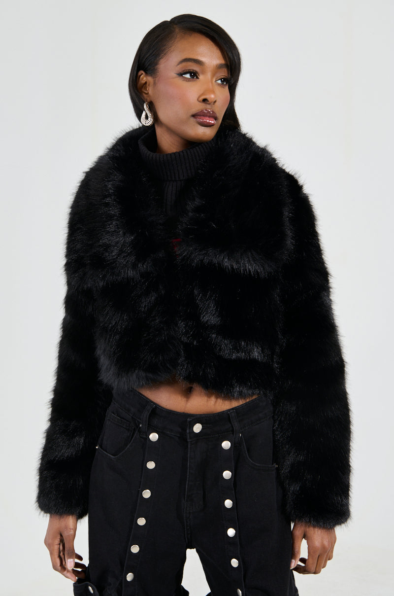 A person wearing the stylish CADENCE FAUX FUR IN BLACK coat and black pants with button details poses against a neutral background, adorned with hoop earrings and exuding a confident expression.