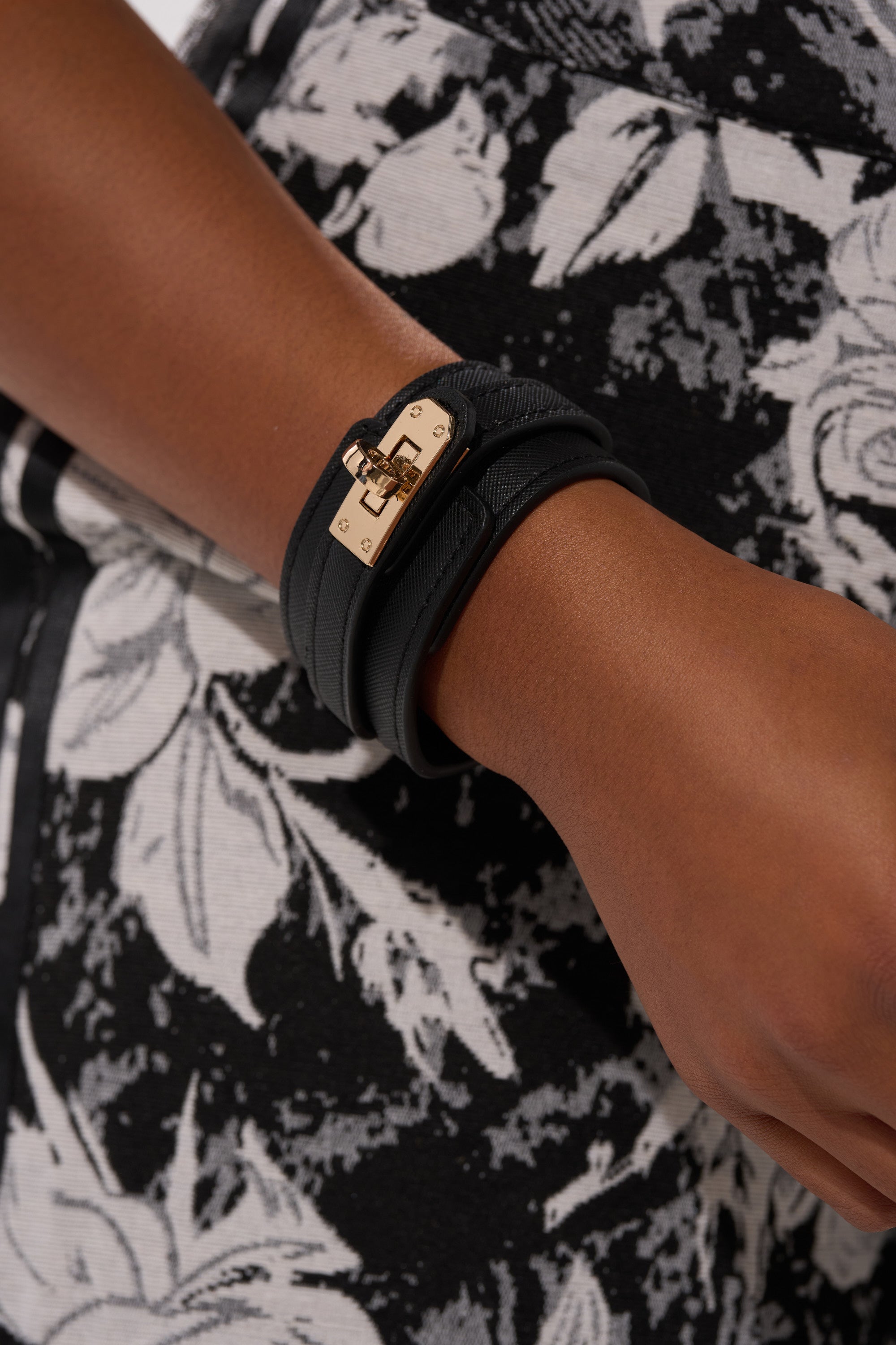 A close-up of a wrist adorned with the DESIGNER BRACELET, featuring a black leather band and gold lock detail, set against black-and-white floral patterned fabric.