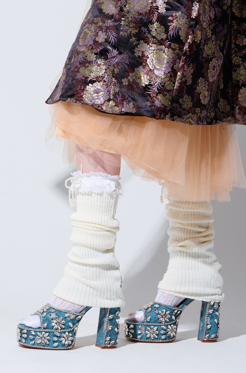 A person stands on a light background wearing a floral-patterned skirt with a peach tulle layer underneath, white leg warmers, and the AZALEA WANG CHARLIZE denim snake skin sandals featuring rhinestones and floral designs.