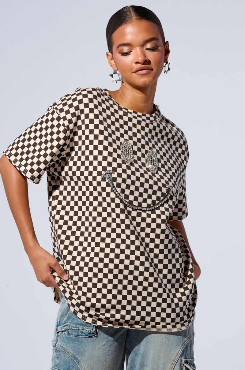 A person with slicked-back hair and statement earrings wears an oversized checkered T-shirt featuring a large smiley face design, known as the Rhinestone Smiley Face Oversized Checkered T-shirt. The cotton-blend fabrication of the shirt complements their light blue jeans, as they stand against a plain light gray background with hands in pockets.