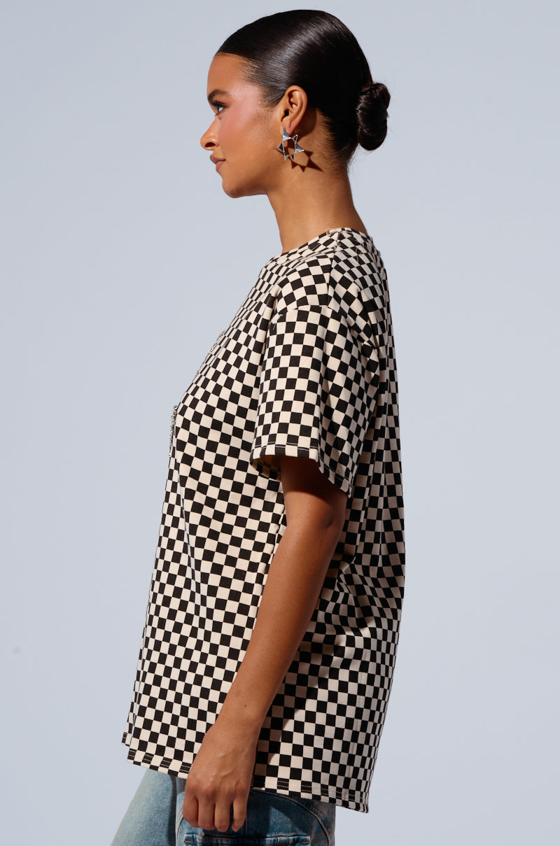 A person with a bun hairstyle is wearing an oversized checkered T-shirt named the RHINESTONE SMILEY FACE OVERSIZED CHECKERED TSHIRT and blue jeans. They are standing sideways against a plain background, showcasing their star-shaped earrings and focused expression. The cotton-blend fabrication of the T-shirt adds comfort to their casual look.