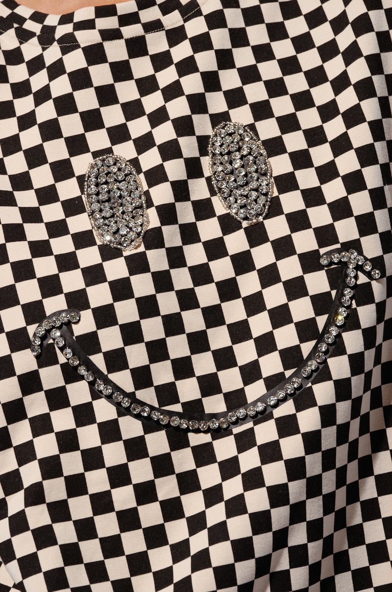 A close-up of the black and white checkered fabric on the RHINESTONE SMILEY FACE OVERSIZED CHECKERED TSHIRT showcases a large smiley face pattern made of glittering rhinestones, featuring two rhinestone eyes and a curved rhinestone mouth on the checkered backdrop.