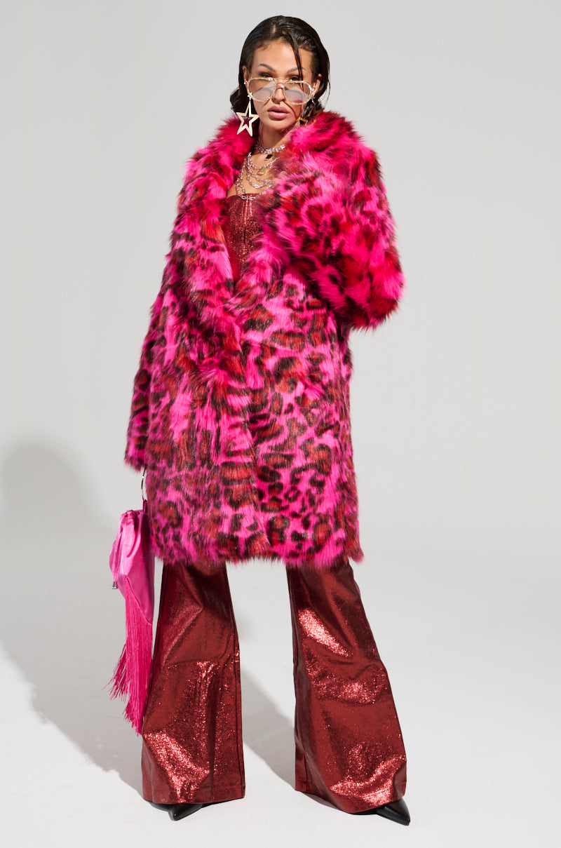 A person poses in the CHEETAH DYNASTY PINK MULTI FAUX FUR JACKET, paired with red metallic flared pants and accessorized with star-shaped earrings. They hold a pink fringed handbag, have their hair styled back, and sport sunglasses against a plain white background, perfectly capturing the essence of bold winter fashion.