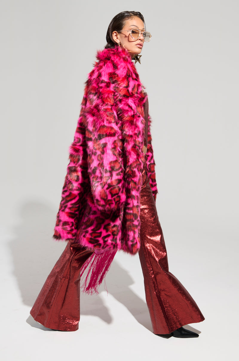 Dressed in shimmering red bell-bottom pants and a matching top, the individual stands out with the CHEETAH DYNASTY PINK MULTI FAUX FUR JACKET. They are accessorized with sunglasses and black pointed shoes, posing against a plain gray background—a true embodiment of bold winter fashion.