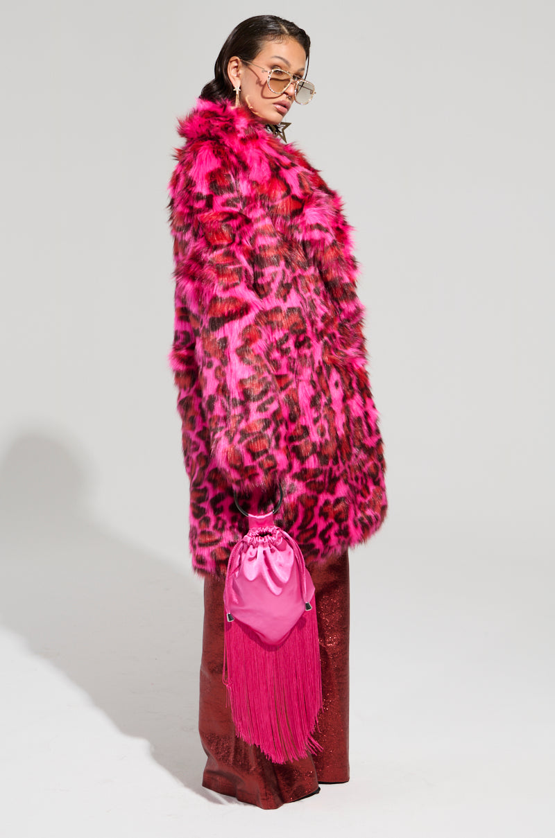 Dressed in the CHEETAH DYNASTY PINK MULTI FAUX FUR JACKET, a person stands confidently against a plain background. They accessorize with sunglasses, gold earrings, and carry a pink tasseled bag. Their deep red textured pants complete the ultimate bold statement look for winter fashion.
