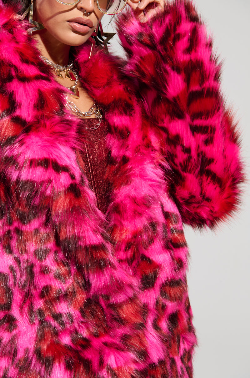 Dressed in the CHEETAH DYNASTY PINK MULTI FAUX FUR JACKET, which showcases a vibrant pink and black cheetah print pattern, the individual exudes confidence. The ensemble is accentuated with oversized round sunglasses and gold chain jewelry, making a bold winter fashion statement against the plain gray backdrop.