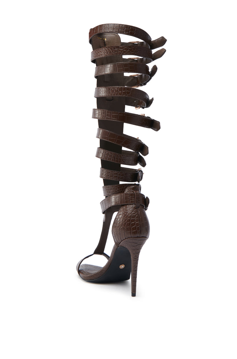 The AZALEA WANG CHERELLI BROWN CROC OVER THE KNEE SANDAL is made from faux croc-embossed leather, with multiple straps extending to the knee, an open toe, a slender stiletto heel, and side buckle closures.