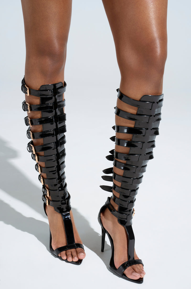 Someone wearing AZALEA WANG CHRELLI BLACK OVER THE KNEE SANDAL—faux patent leather, gladiator-style with adjustable buckle straps—stands on a light background.