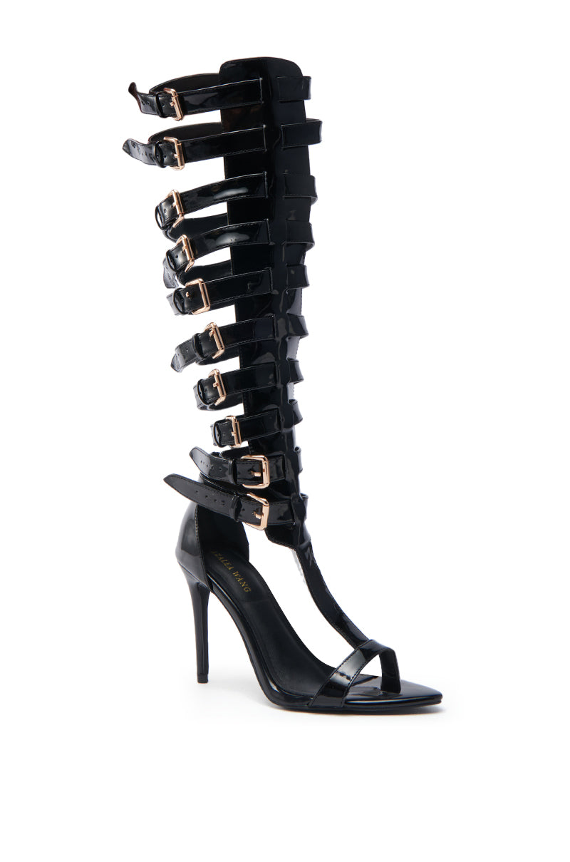 The AZALEA WANG CHRELLI BLACK OVER THE KNEE SANDAL features a faux patent leather finish, adjustable buckle straps, shiny gold calf accents, open toe design, and high stiletto heel.
