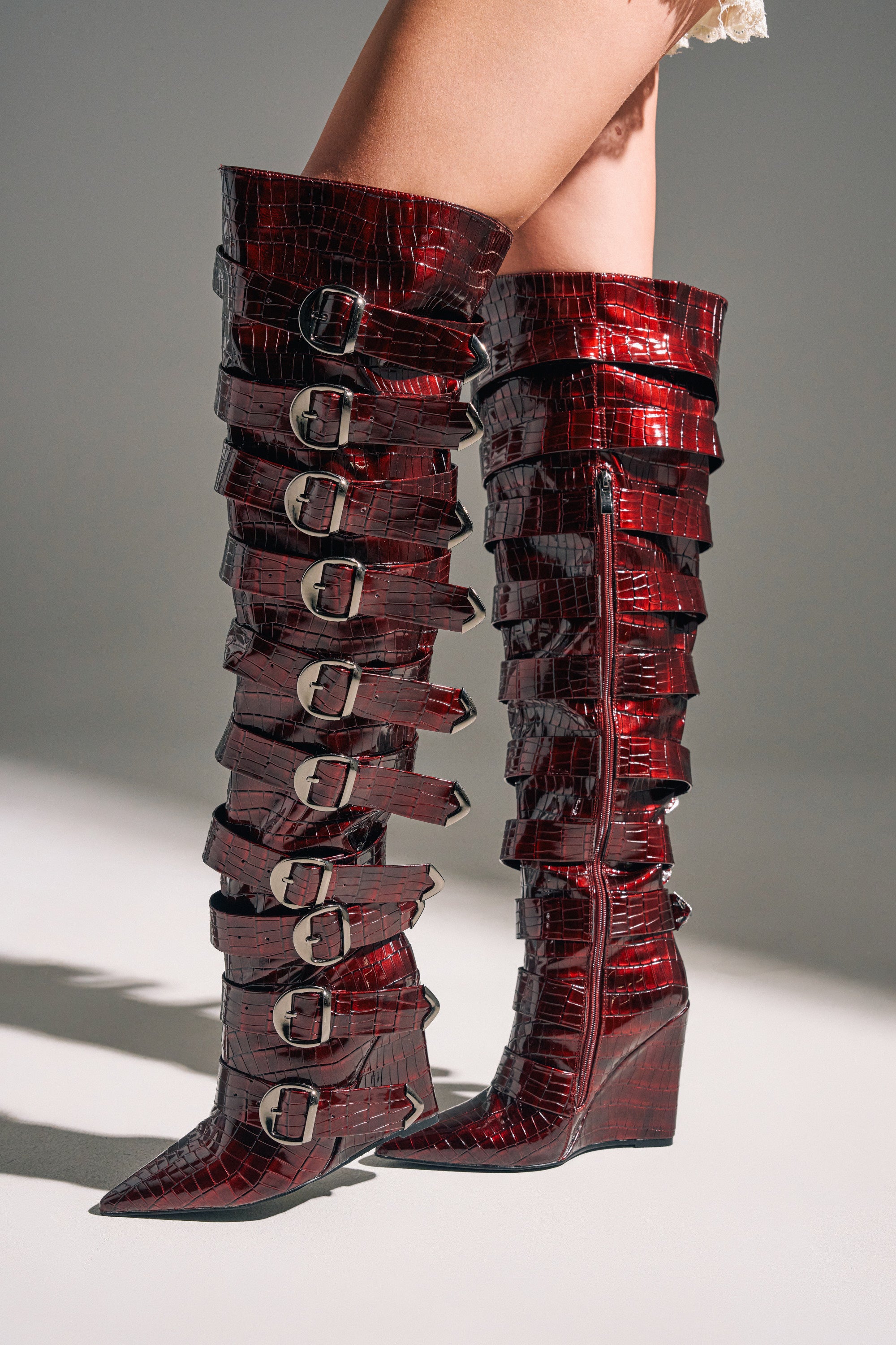 Someone stands on a light surface wearing the AZALEA WANG CINZIA RED WEDGE BOOT WITH BUCKLE DETAIL—tall, shiny red boots featuring faux crocodile leather, multiple silver buckles, and pointed toes.