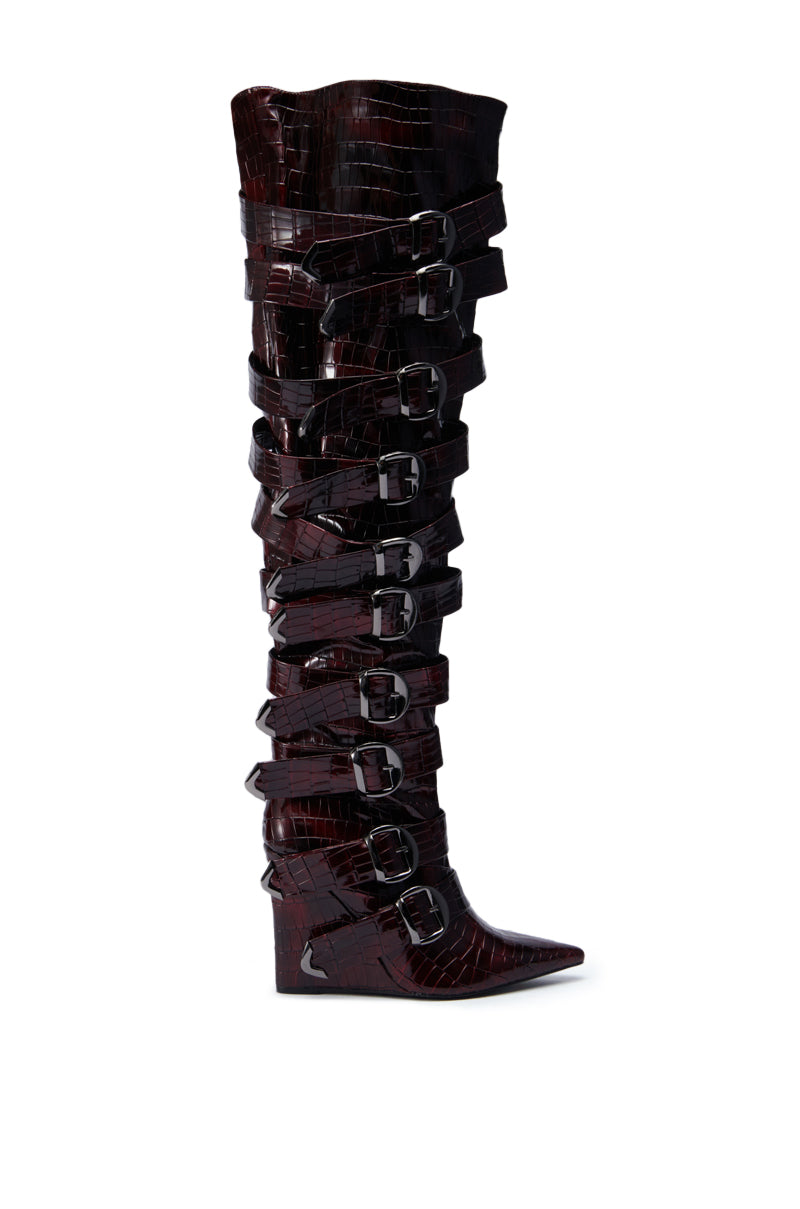 The AZALEA WANG CINZIA RED WEDGE BOOT WITH BUCKLE DETAIL is a tall, pointed-toe wedge boot in deep red faux crocodile leather, featuring multiple silver buckles and straps with a glossy finish and striking wedge heel.