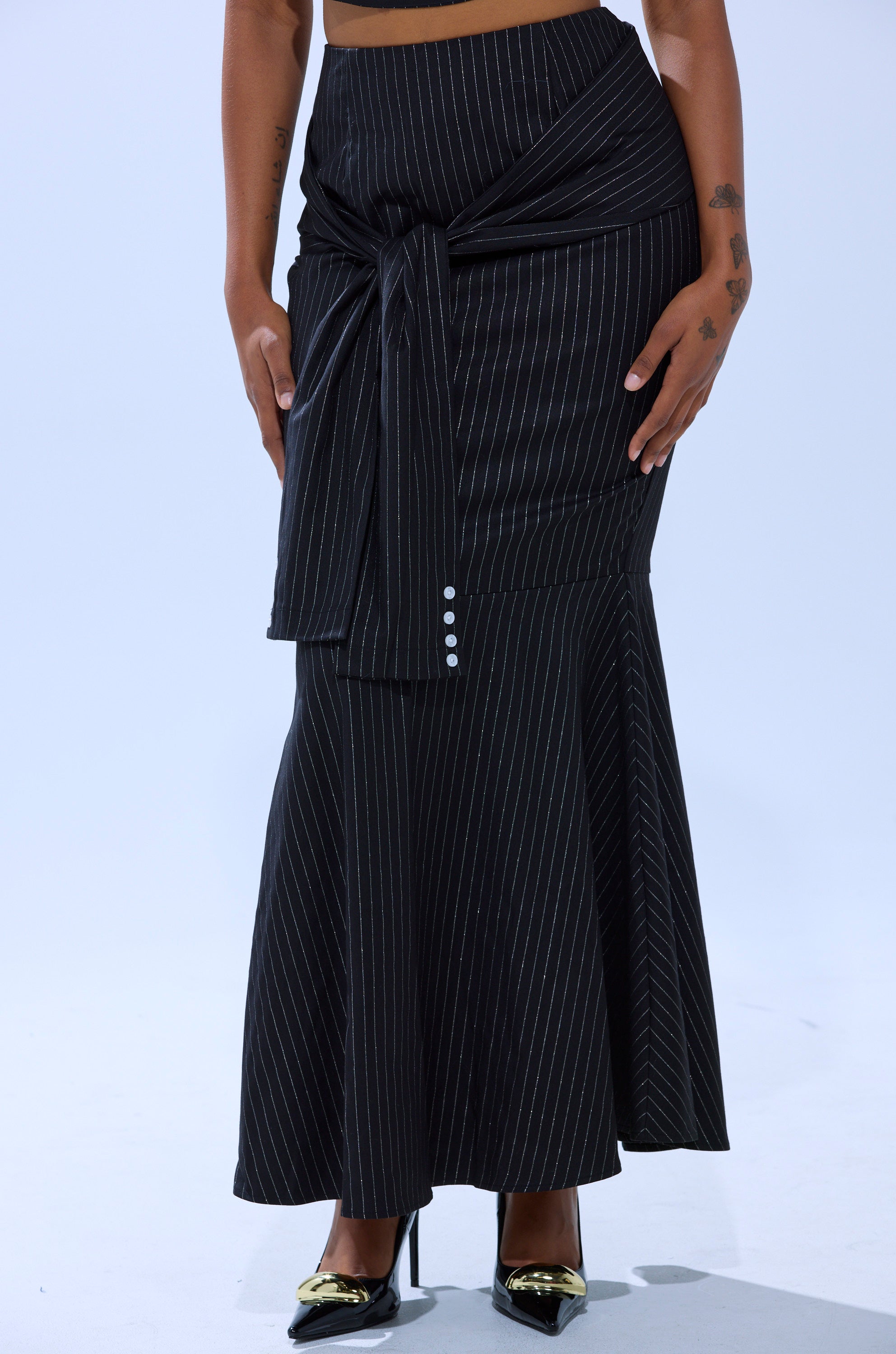 Wearing the BUSINESS ONLY PINSTRIPE MAXI SKIRT with a front tie and decorative buttons, paired with black high heels featuring metallic gold pointed toes.
