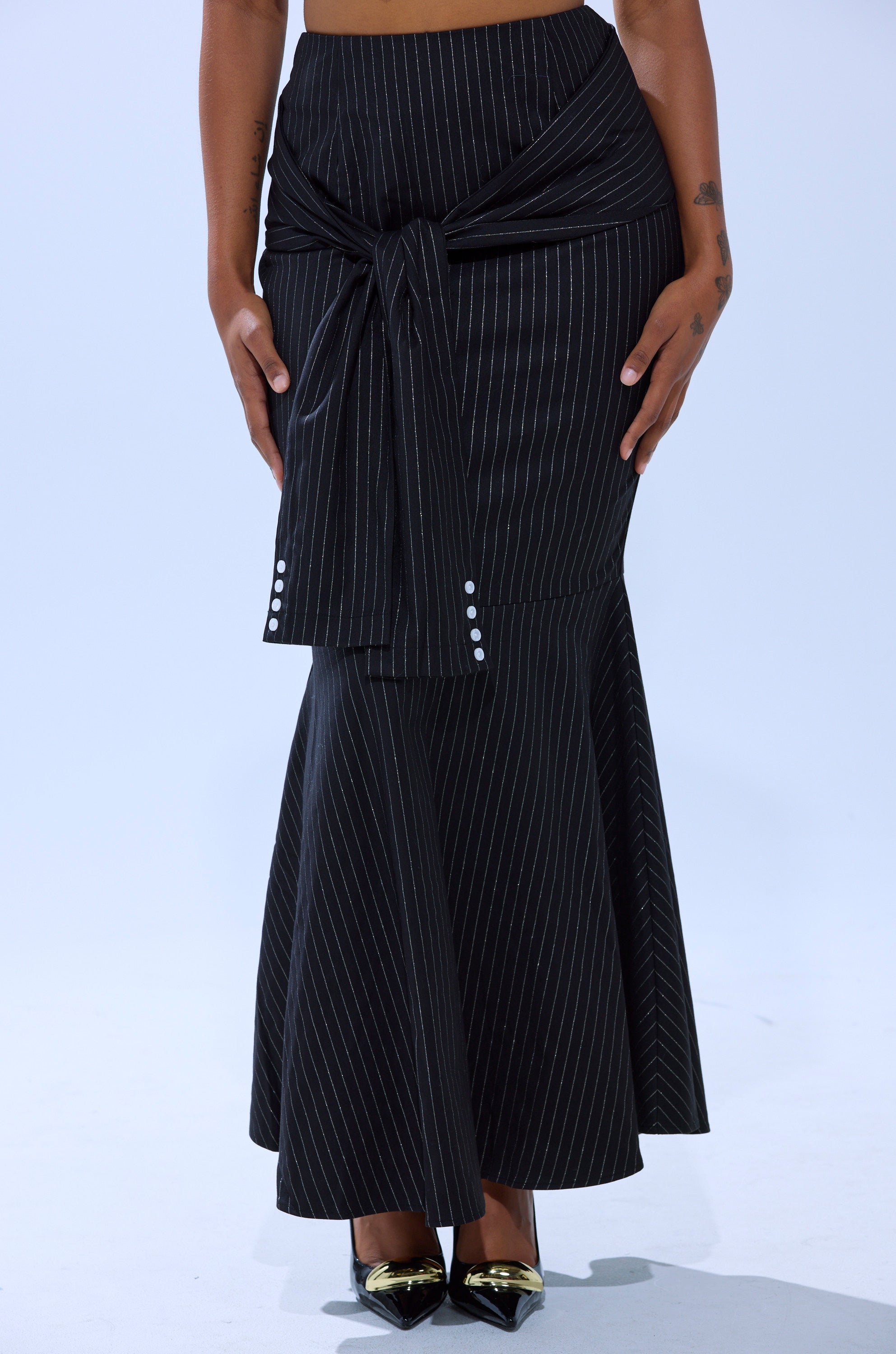 Someone wears the BUSINESS ONLY PINSTRIPE MAXI SKIRT—a black, pinstripe maxi with a flared mermaid silhouette, wide tied sash waist, and decorative buttons—paired with black pointed-toe heels with metallic gold caps.