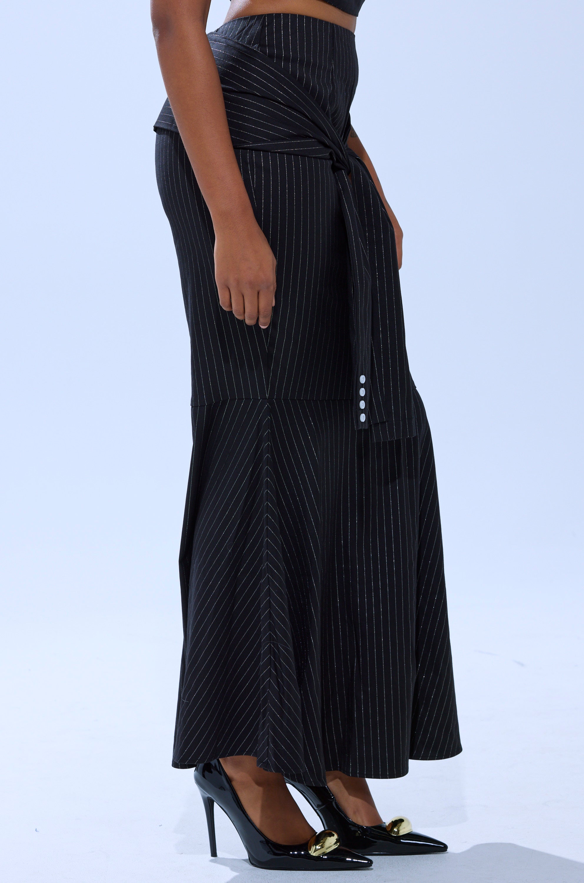 A person wears the BUSINESS ONLY PINSTRIPE MAXI SKIRT, a black high-waisted mermaid silhouette with front tie, paired with black heels featuring gold accents, standing against a light background.