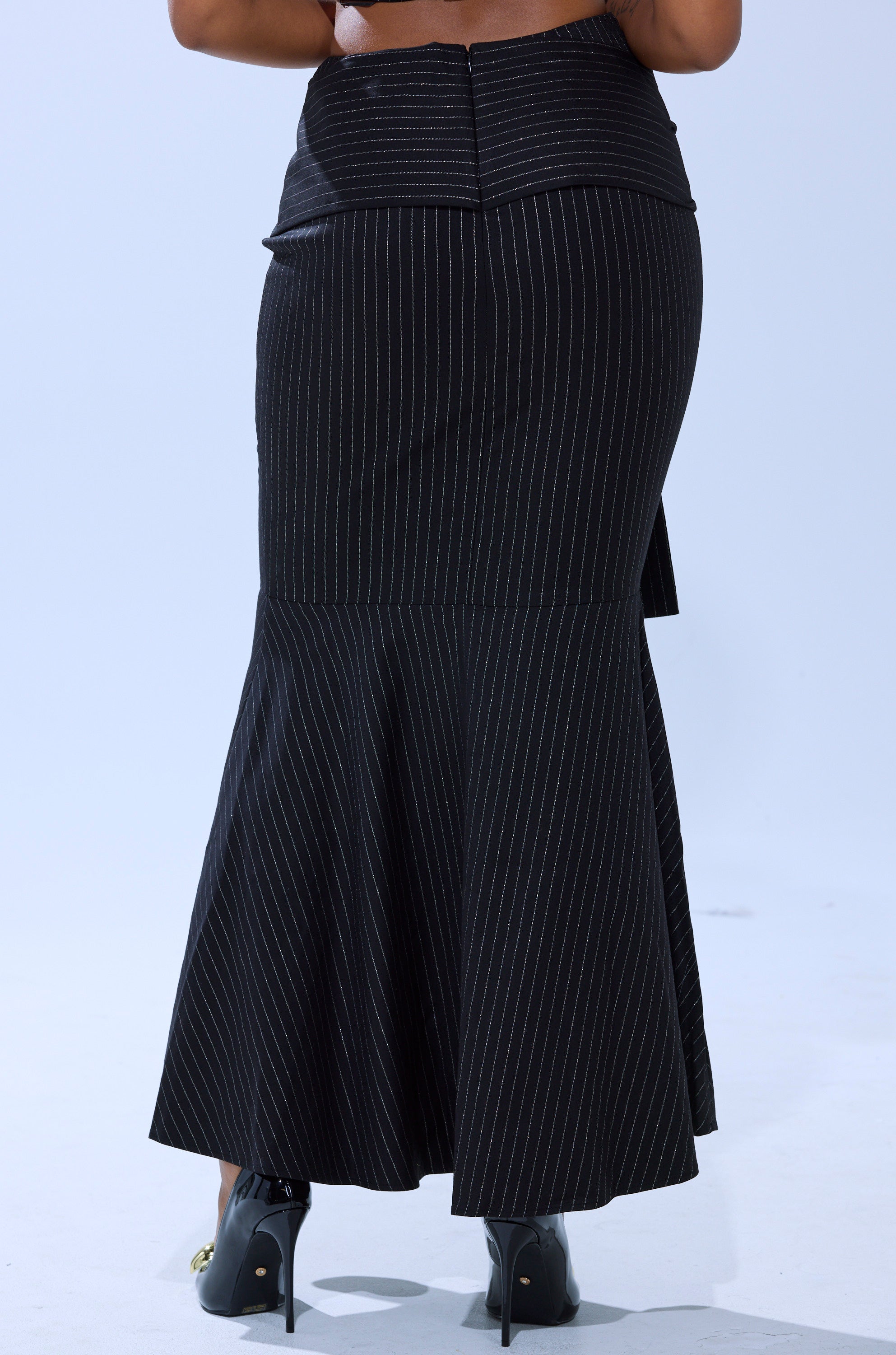 Seen from the back against a plain light background, a person wears the BUSINESS ONLY PINSTRIPE MAXI SKIRT—high-waisted, black, with a mermaid silhouette and flared hem—paired with black high-heeled shoes.