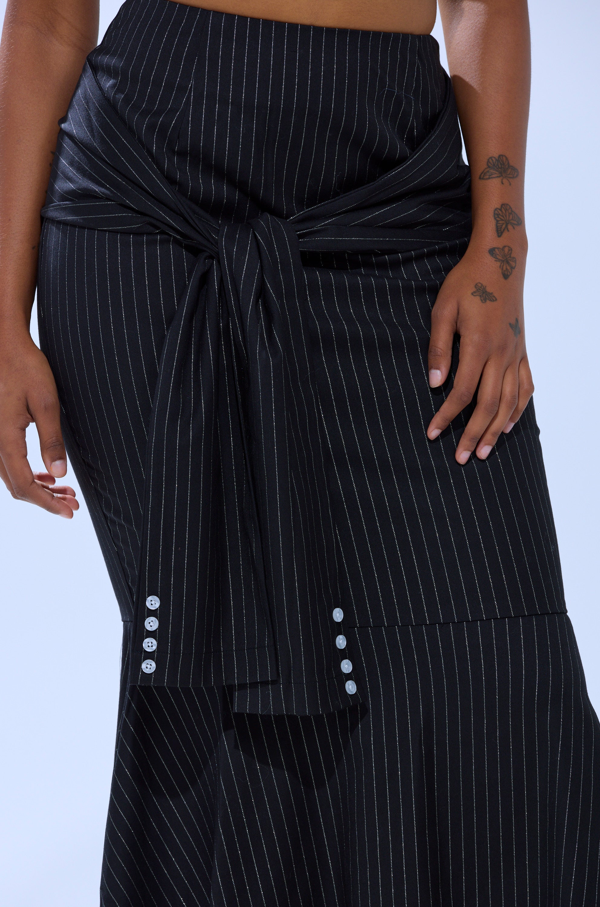 Wearing the BUSINESS ONLY PINSTRIPE MAXI SKIRT—black with pinstripes, a large front tie, and decorative buttons—a person with butterfly tattoos on their arm stands with their hand resting at their side.