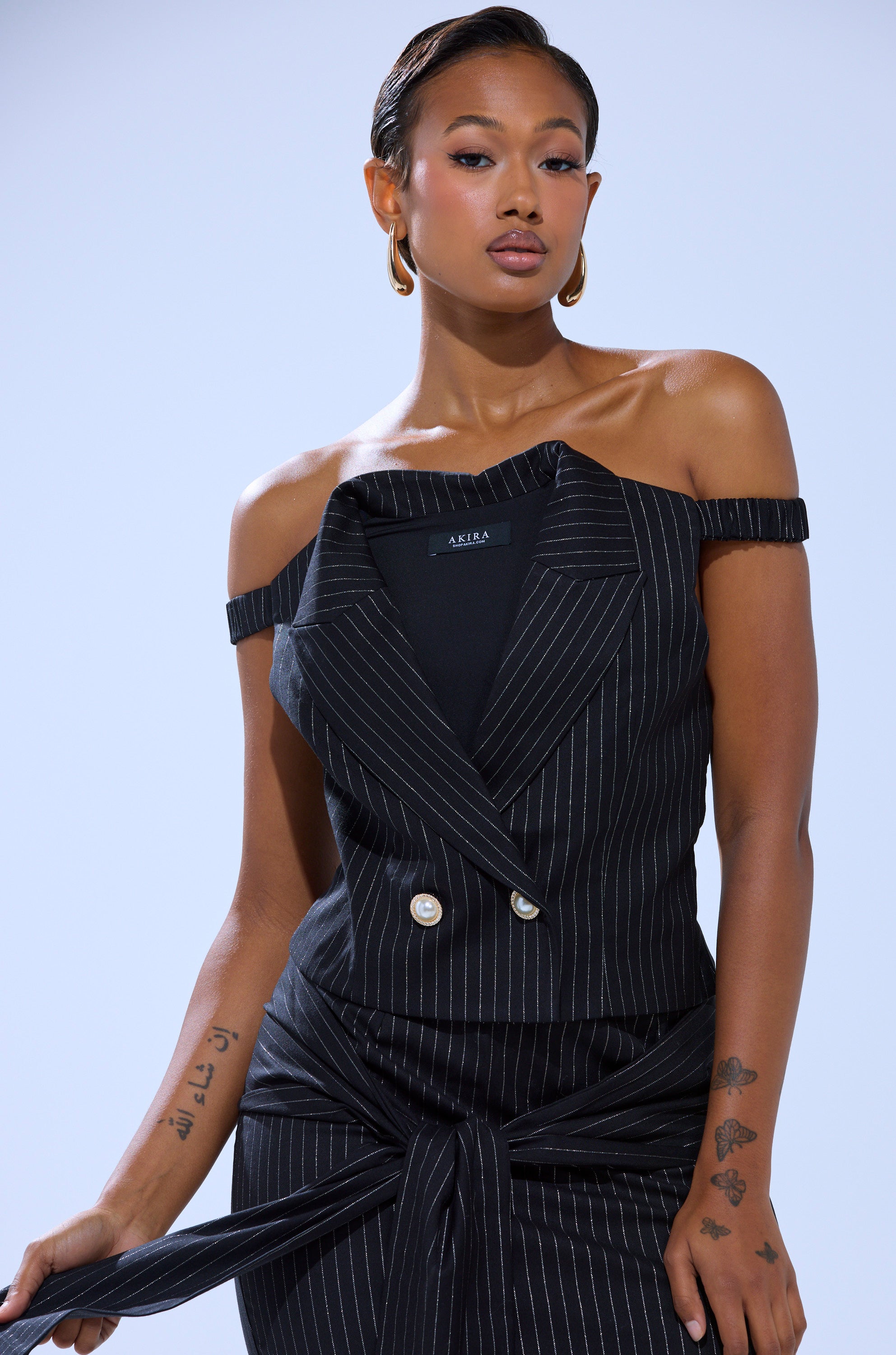 A woman confidently poses in the BUSINESS ONLY PINSTRIPE CORSET from AKIRA Label, styled with gold hoop earrings, slicked-back hair, and visible left arm tattoos. The outfit features black pinstripes and gold buttons against a plain light background.