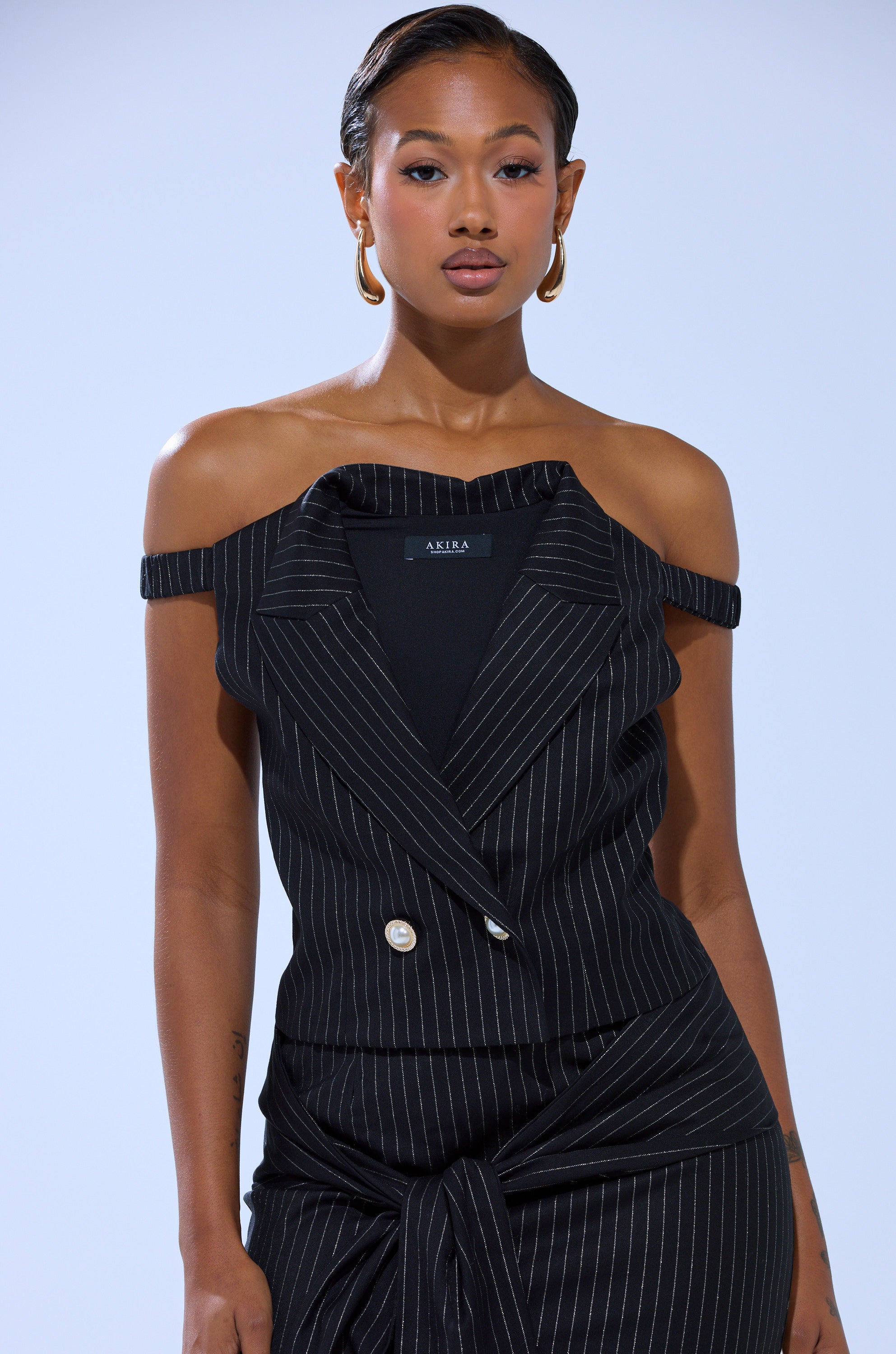 A woman wearing the AKIRA Label BUSINESS ONLY PINSTRIPE CORSET with gold hoop earrings stands against a light blue background.