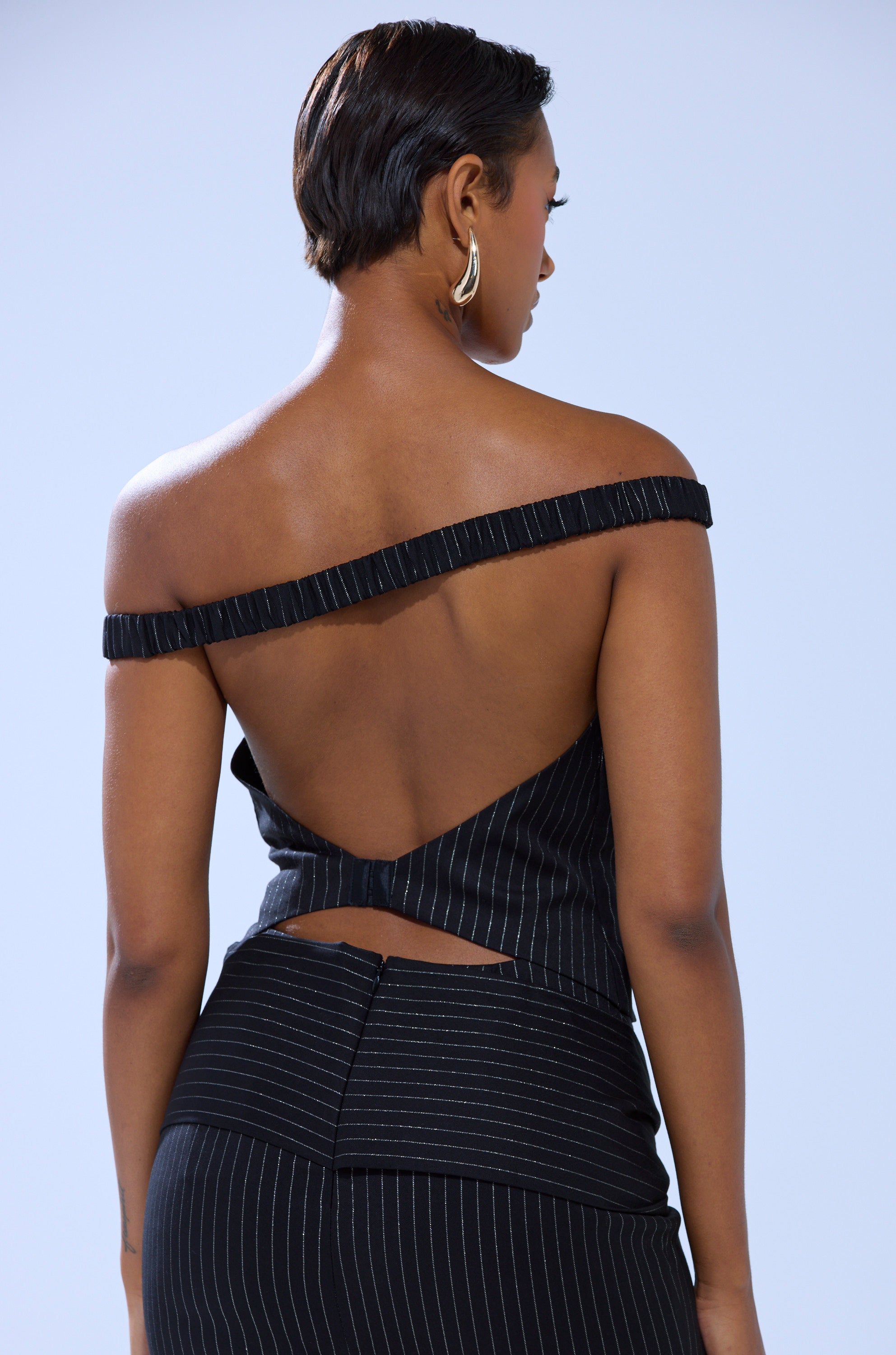A woman with short hair wears the BUSINESS ONLY PINSTRIPE CORSET, a black, strapless, backless top paired with matching pinstripe pants, standing with her back to the camera against a light background.