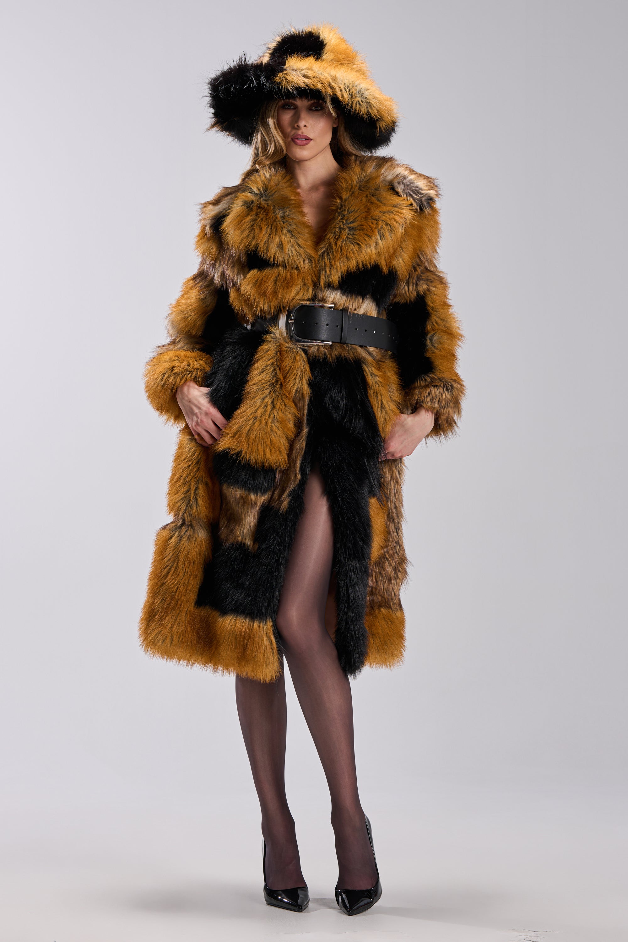 A woman stands confidently in the COSMO PATCHWORK FAUX FUR jacket and matching wide-brim hat, both with orange, black, and brown patterns. She styles them with a wide black belt, sheer tights, and heels against a plain background.