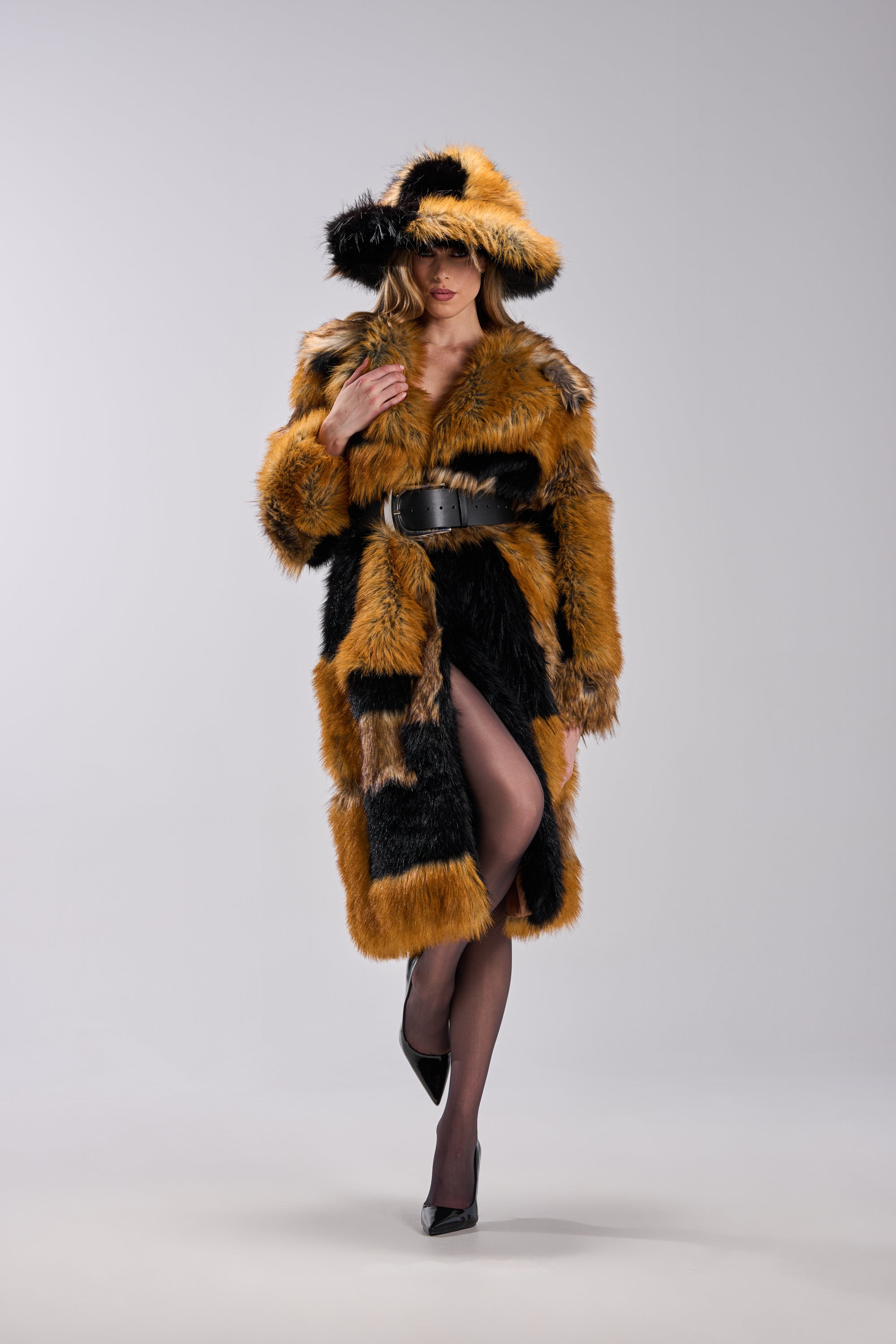 A woman models the COSMO PATCHWORK FAUX FUR jacket and matching hat, both with striking orange and black patches. She accessorizes with a black belt, sheer stockings, and heels, her face partially obscured by the hat against a plain backdrop.