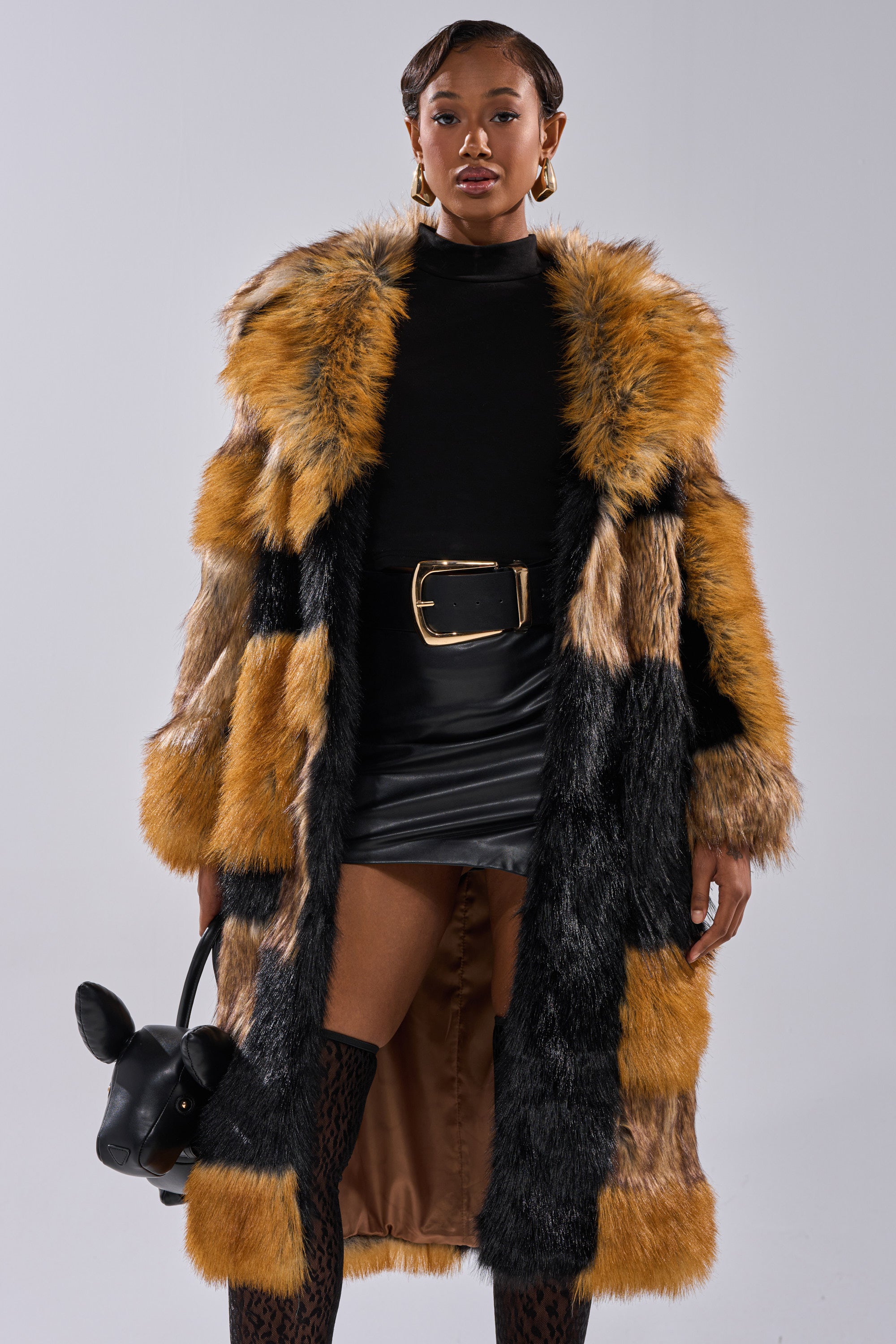 A stylish woman wears the COSMO PATCHWORK FAUX FUR coat over a black turtleneck and leather mini skirt, accessorized with a wide belt, gold hoop earrings, and holding a black cat-shaped handbag.