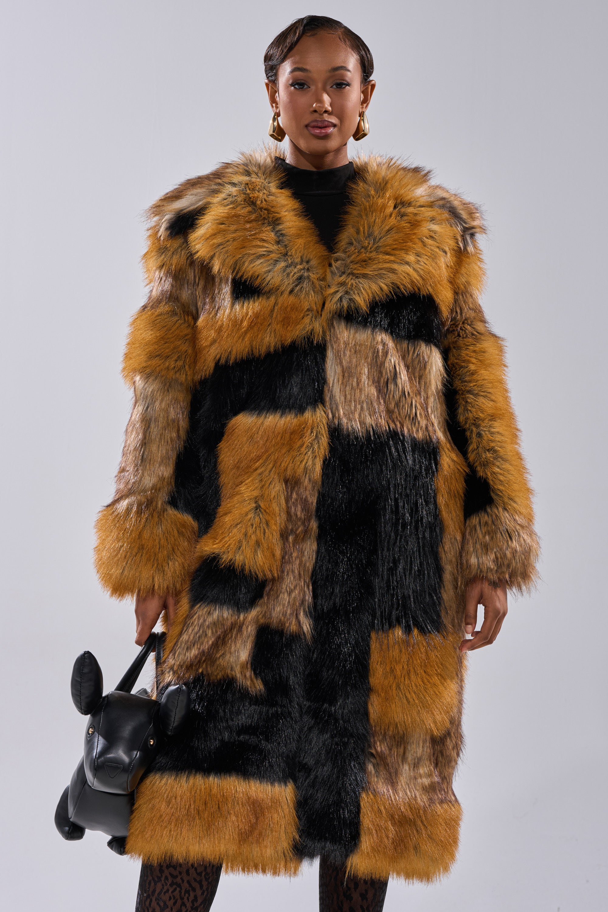 A woman poses against a plain background wearing the COSMO PATCHWORK FAUX FUR jacket in black, brown, and tan. She accessorizes with gold hoop earrings and carries a black dog-shaped handbag.