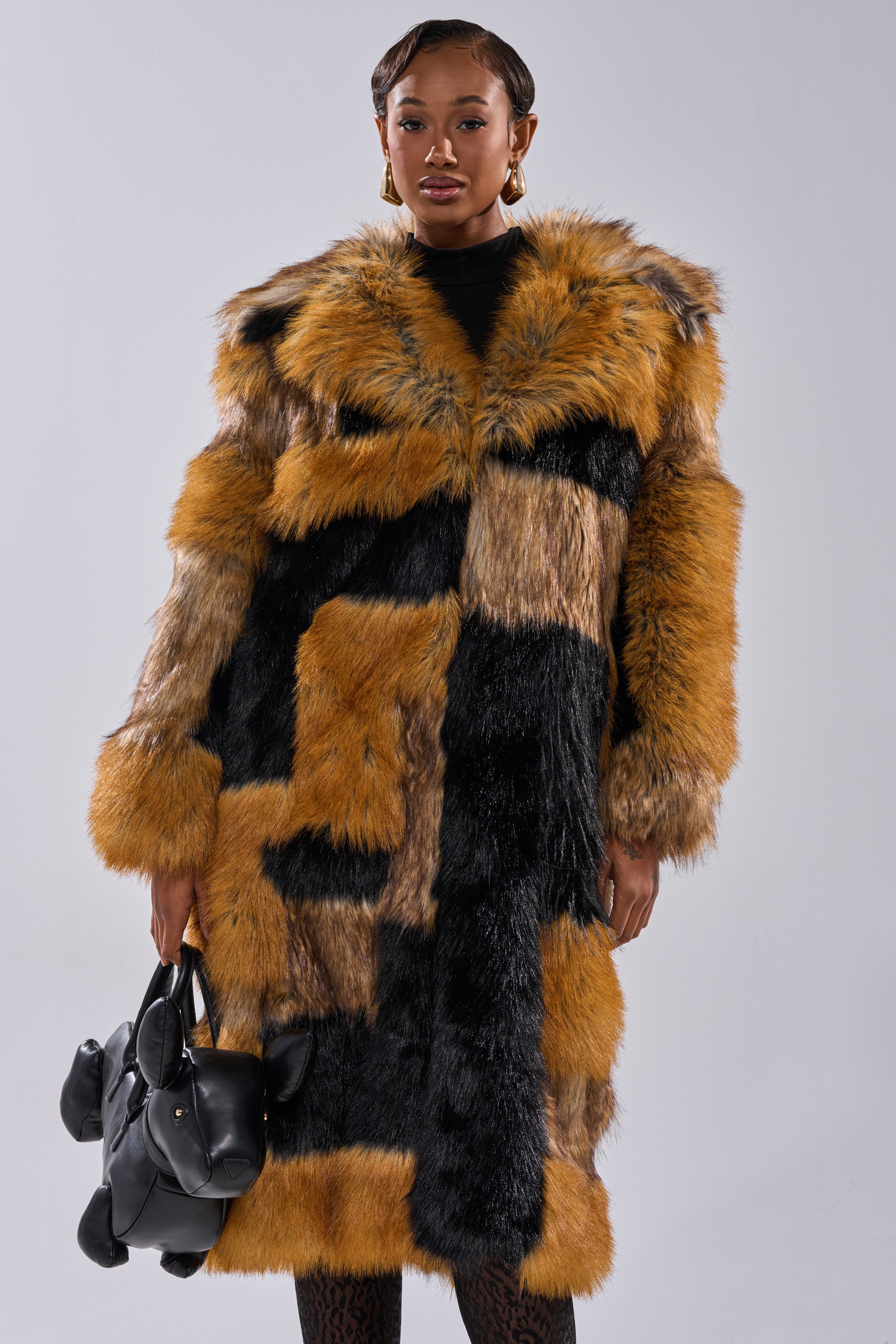 A woman models the COSMO PATCHWORK FAUX FUR coat in brown, black, and golden tones, standing against a plain background and holding a dog-shaped black handbag.