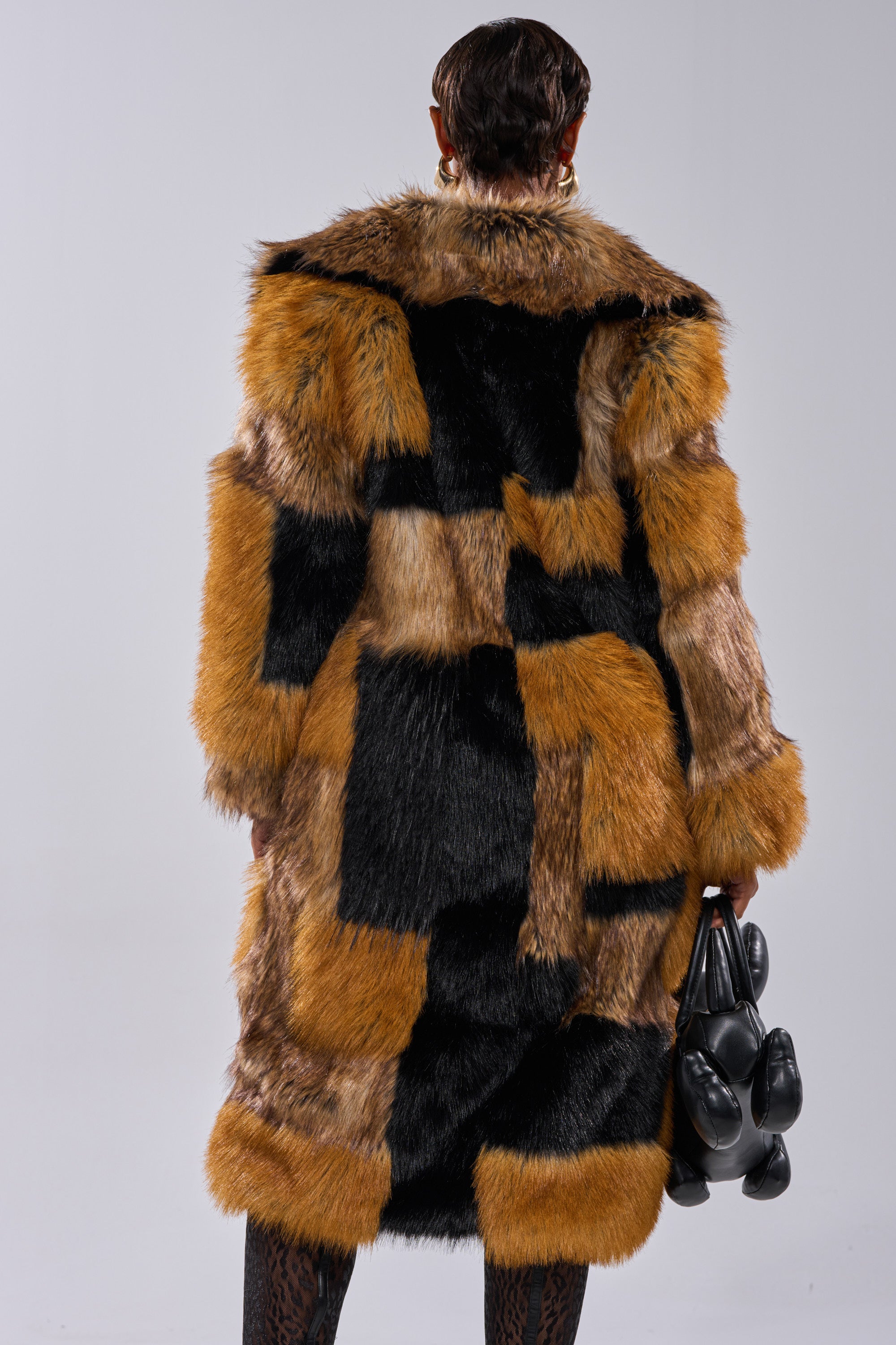 A person stands facing away against a plain gray background, wearing the COSMO PATCHWORK FAUX FUR coat in brown, black, and tan shades, and holding a black abstract handbag in their right hand.