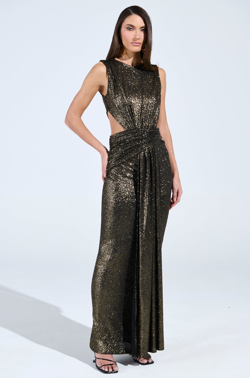 A person poses in a shimmering gold gown with glitter detailing and side cutouts, similar to the CROSS MY HEART MAXI DRESS WITH CUTOUT. The cinched waist and flowing skirt pair perfectly with their black strappy heels, as long hair cascades against the plain background.