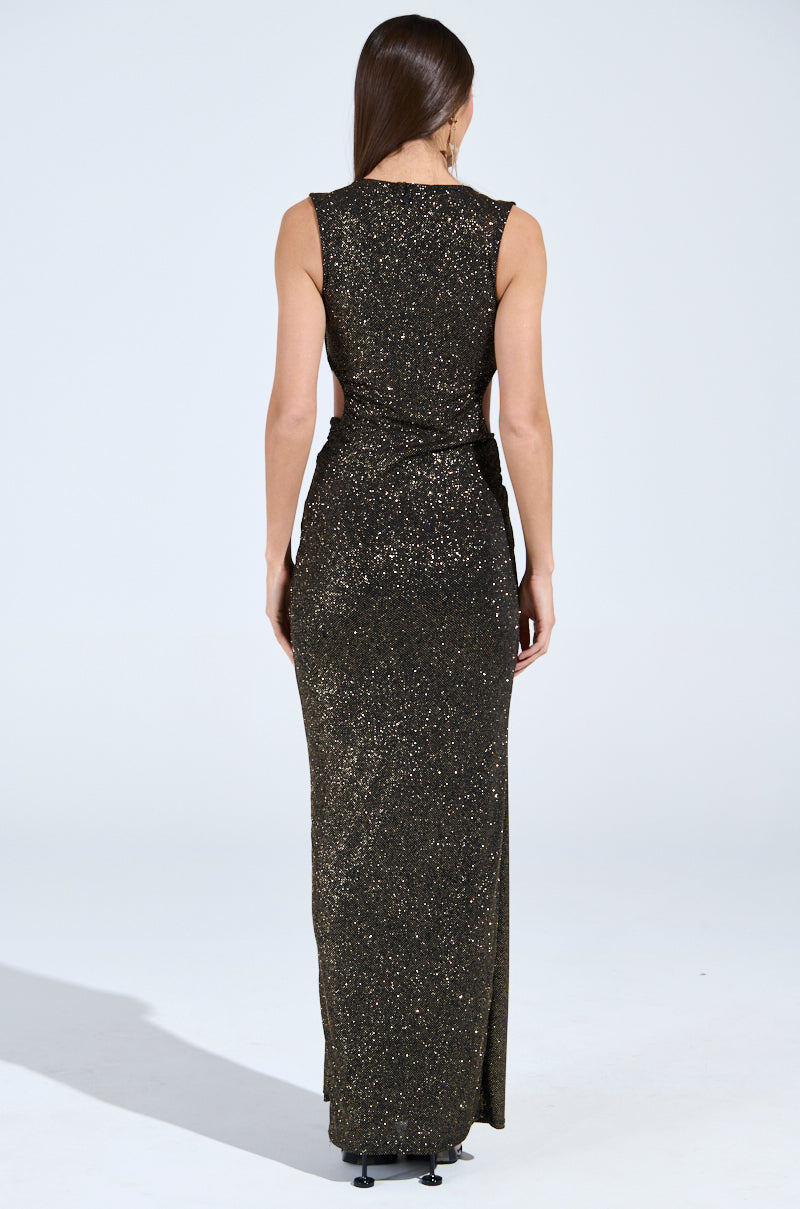 A woman wearing the CROSS MY HEART MAXI DRESS WITH CUTOUT poses with her back to the camera, highlighting a full-length, sleeveless gown embellished with gold glitter. Her long hair flows down her back in front of a plain white background.