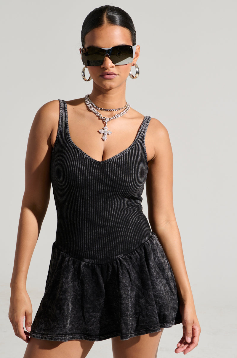 A woman wearing the HELLO SUMMER KNIT MINI DRESS in a sleek, black ribbed fabric stands against a plain background. She accessorizes with large hoop earrings, layered necklaces, and dark sunglasses. Her hair is pulled back neatly.