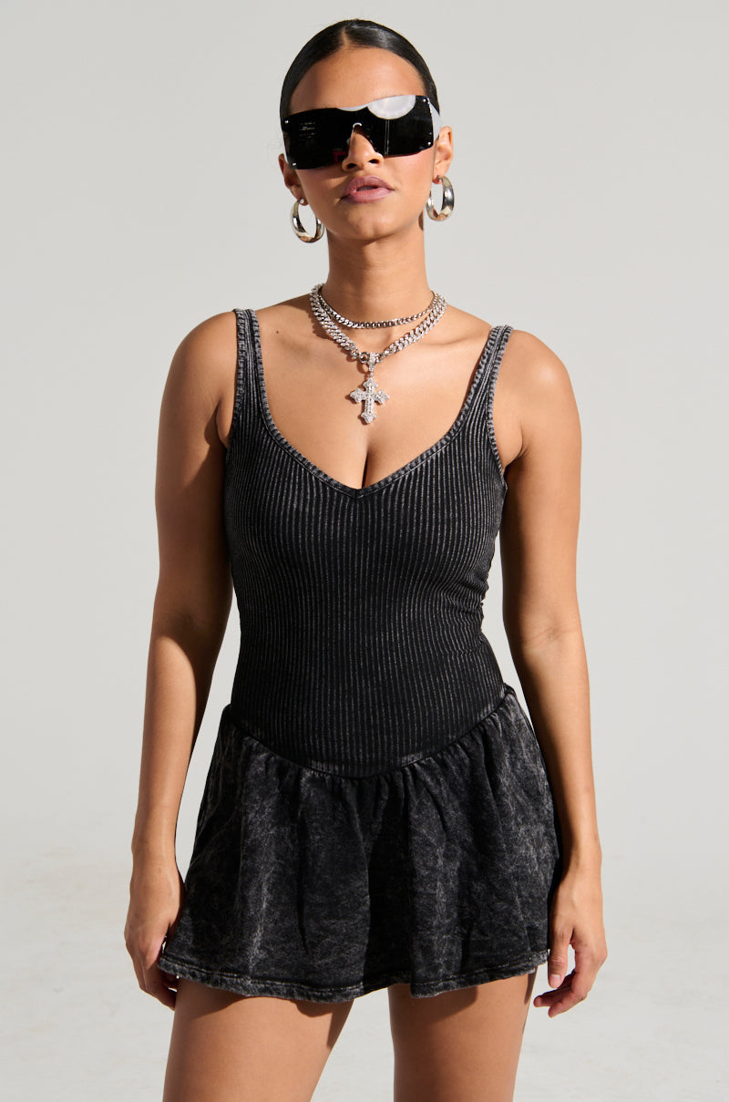 A woman confidently stands wearing large black sunglasses, hoop earrings, and a silver necklace. She is dressed in the HELLO SUMMER KNIT MINI DRESS, featuring a ribbed knit bodice and a short, dark gray tonal flared skirt. Her hair is pulled back, exuding a fashionable and fierce look against a plain background.